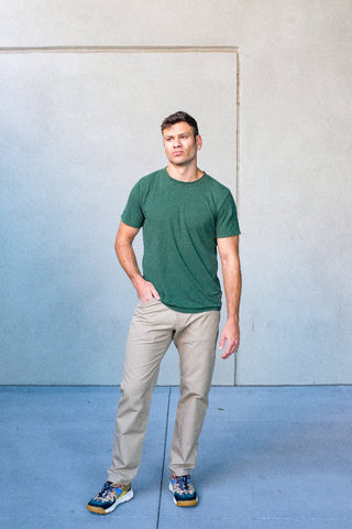 Man wearing a green t-shirt and beige pants standing against a light blue wall.
